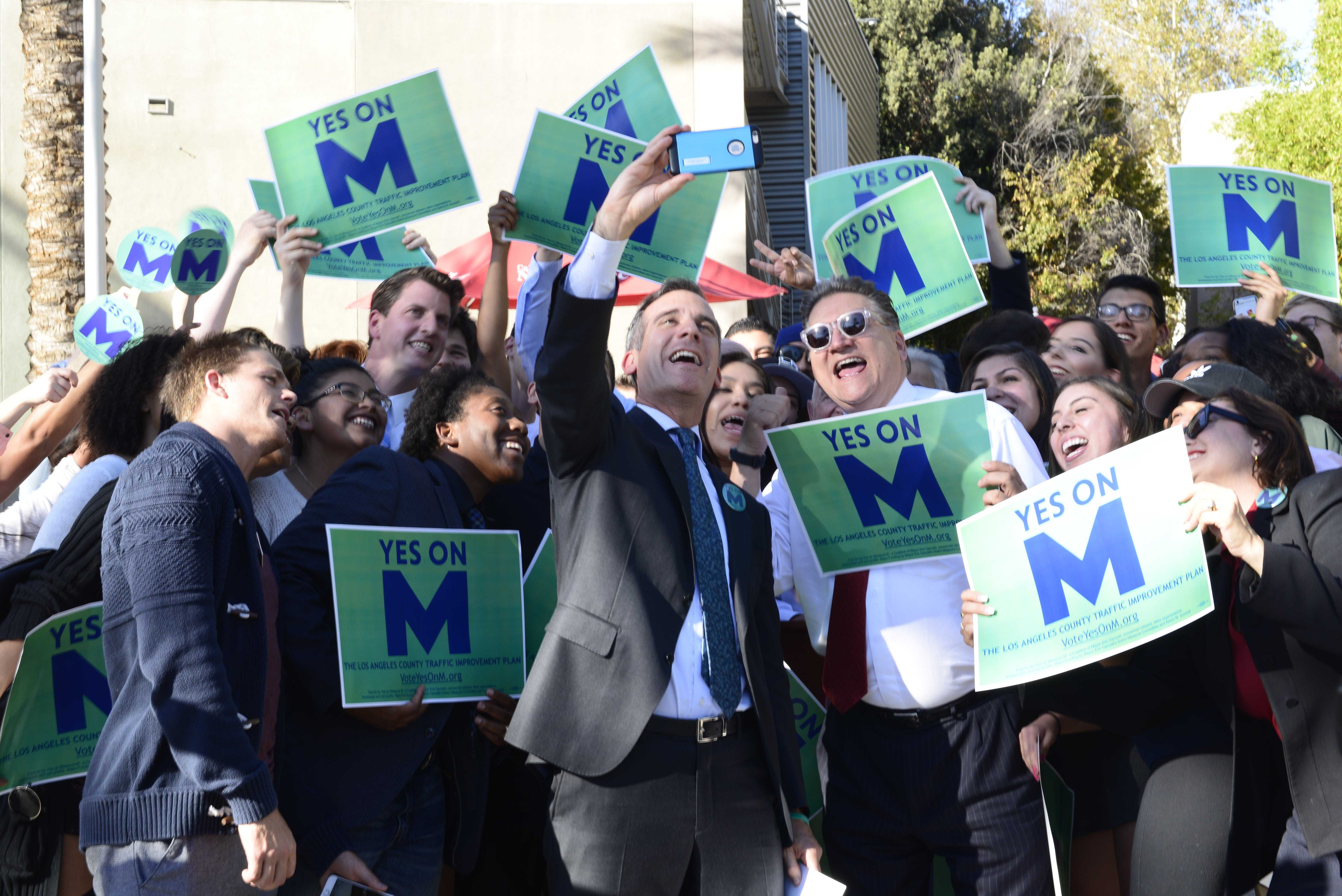 Mayor shown taking picture with people holding signs that read, Yes on M