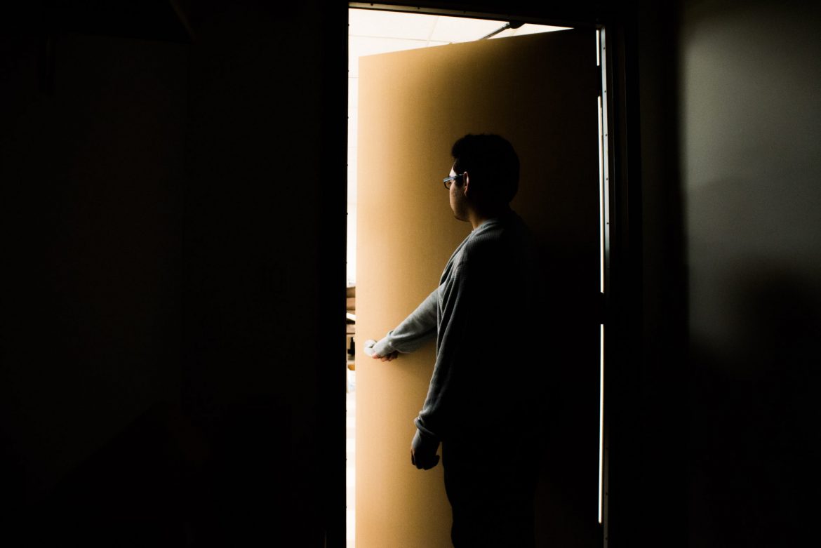 Man pictured in a dark room opening a door into a lit room