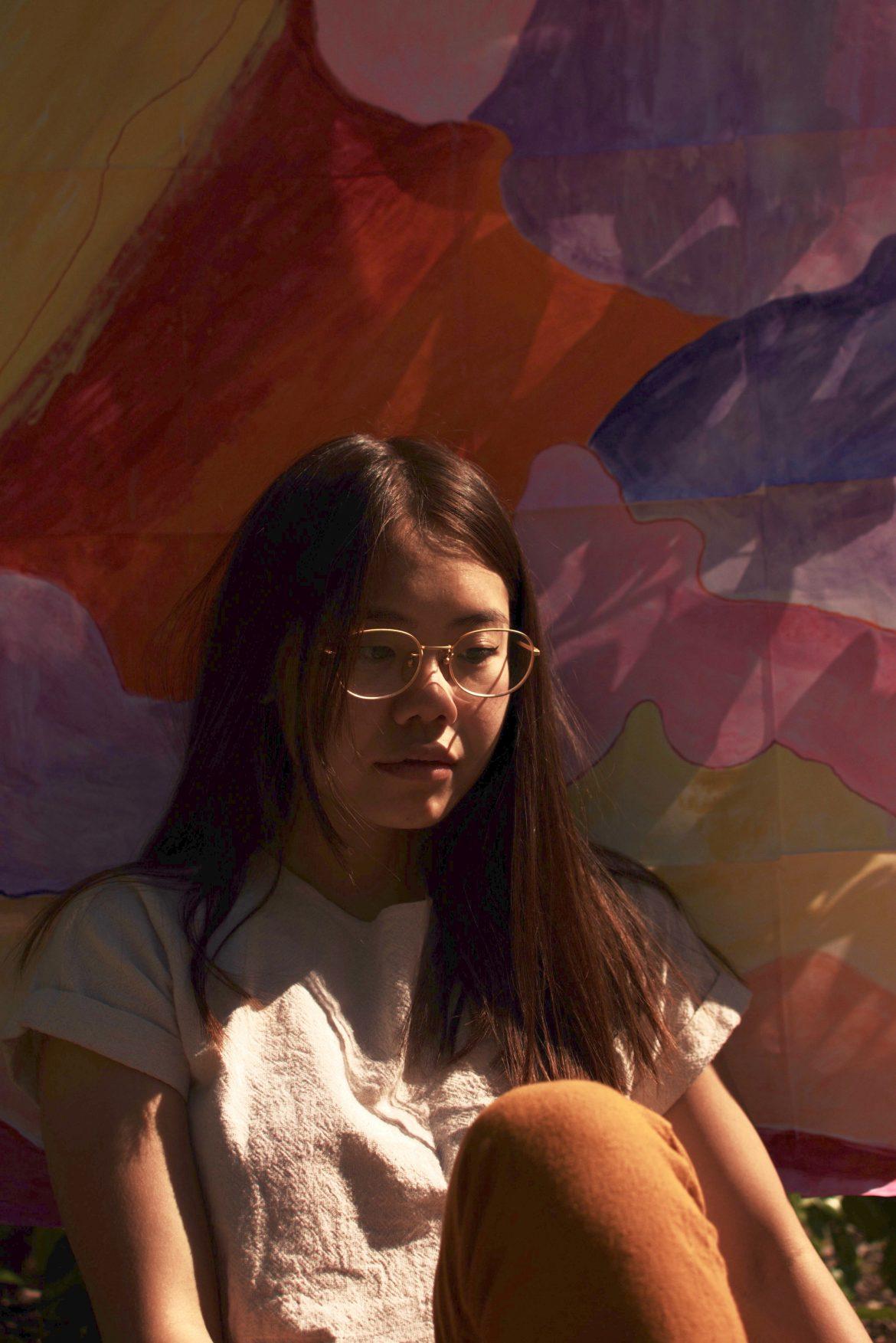 Trina, a young woman with black hair and glasses sits in front of a colorful abstract painting