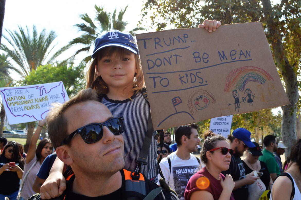 Little girl sits on her dads shoulders at protest with a sign that says, Trump dont be mean to kids