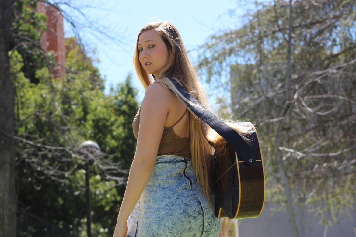 woman with guitar strapped around her back looks seriously into the camera