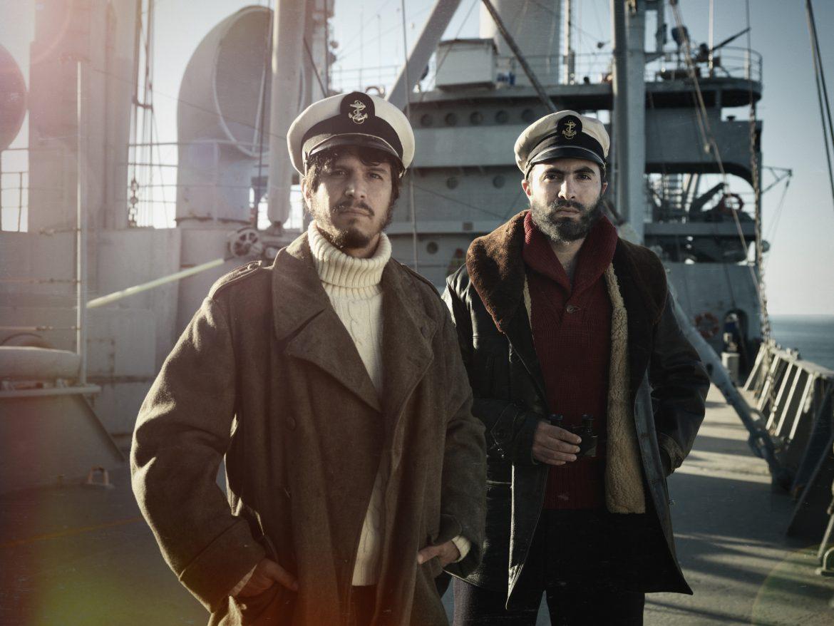 two men dressed warmly standing on the deck of a boat with sailor hats on