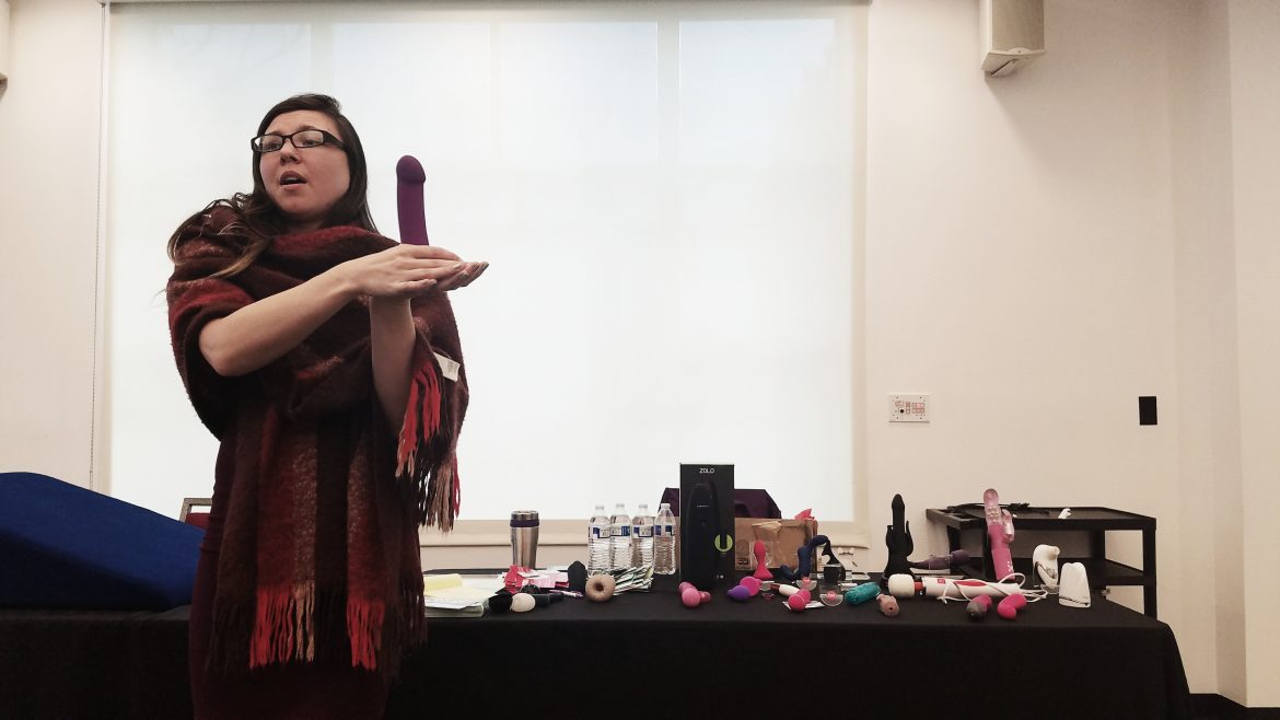 woman+wrapped+in+scarf+holds+purple+dildo+in+palm+while+speaking+to+an+audience