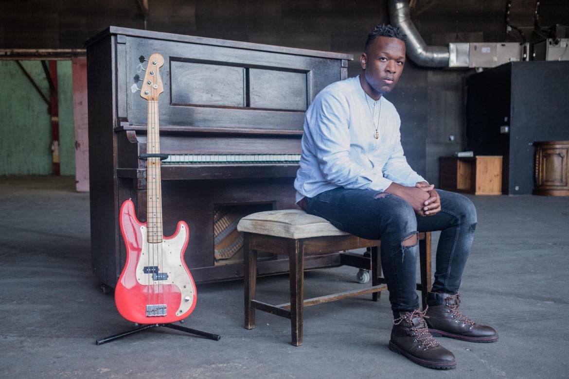 A male African American musician sitting on a chair in front of a piano and red electtrical guitar by the side.