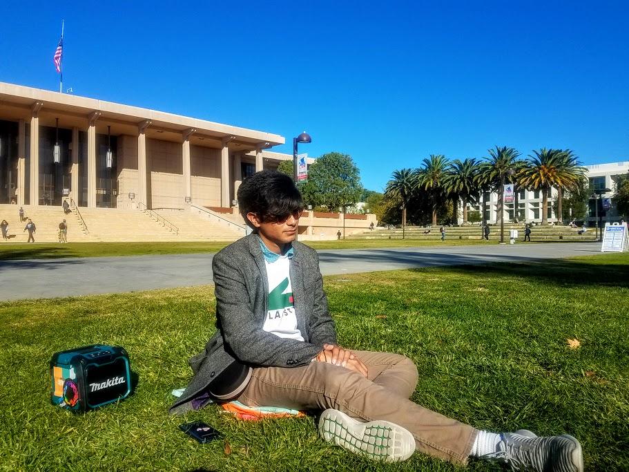 A male student sitting on the oviatt lawn enjoying the sunshine and having a colorful bluetooth speaker on the side.