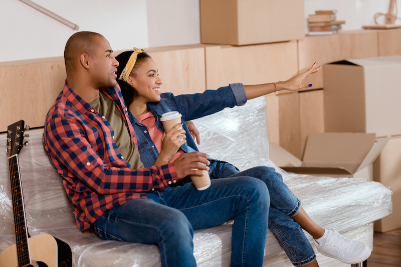 young male and female couple sitting on a new couch surrounded by moving boxes