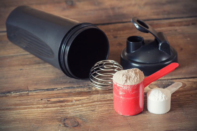 Whey+protein+powder+in+scoop+and+plastic+shaker+on+wooden+background