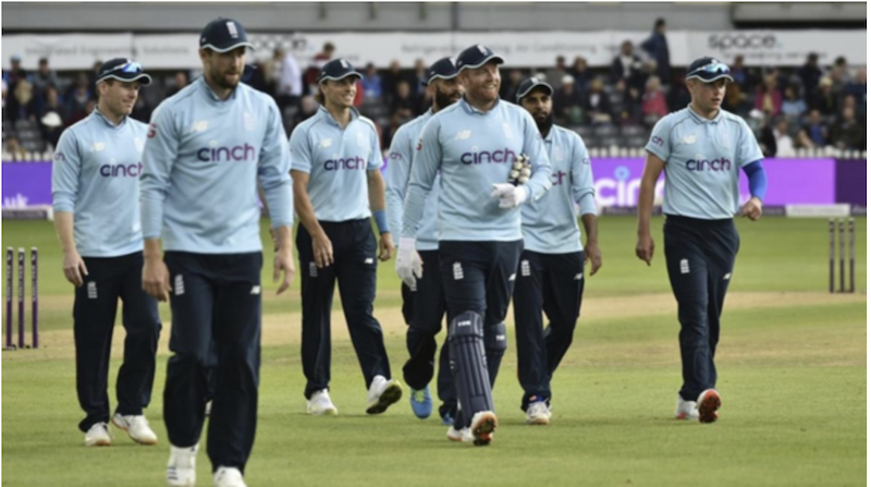 Cricket players in light blue shirts and dark pants and hats walk across a field