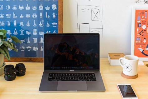 black laptop, camera lenses, phone and coffee mug on wooden desk