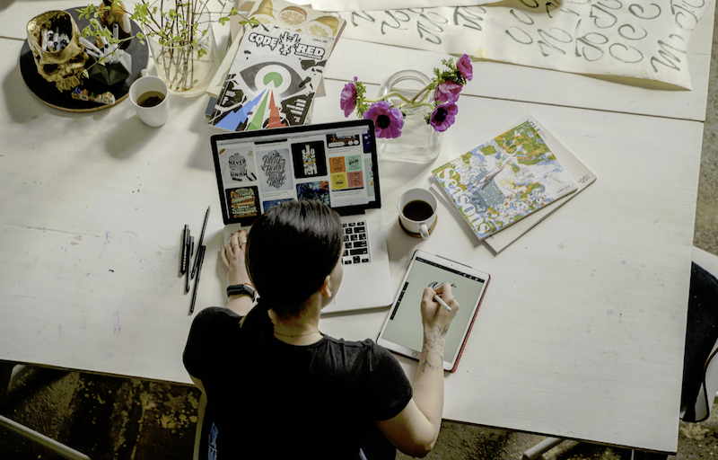 A picture of a woman with her iPad and computer. She is drawing on her iPad. There are some books, flower and coffee on the table as well.