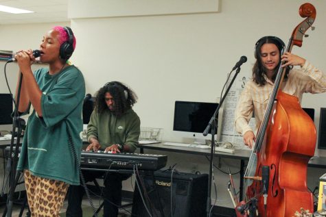 three people doing music in a room. All of them are very focus and doing something different. The girl is singing, one of the boys is on the piano, and the other one playing a cello