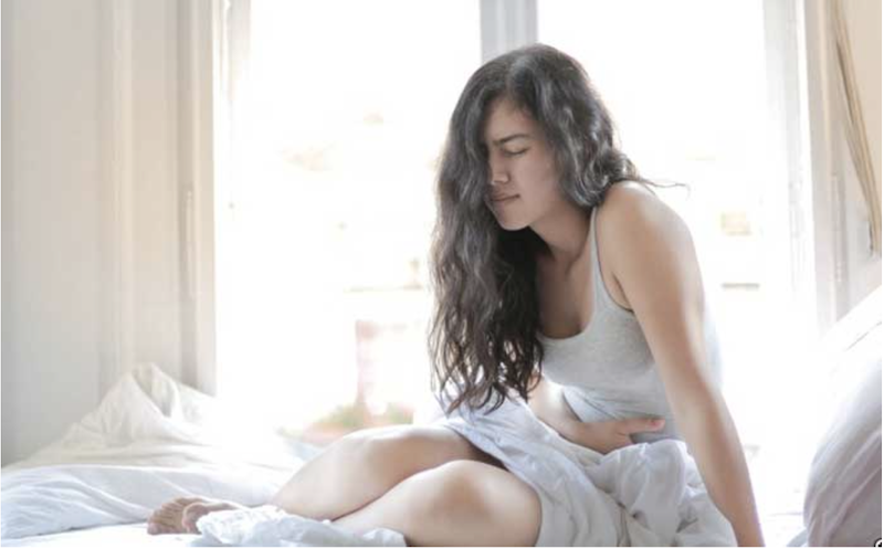 Woman with long dark hair sitting on bed holding her stomach