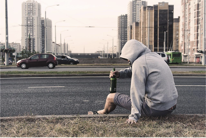 man+in+gray+hoodie+sitting+by+road+and+holding+liquor+botle