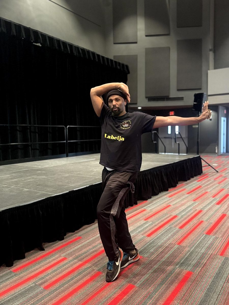 Man wearing all black, has one arm out to the side and the other on top of his head